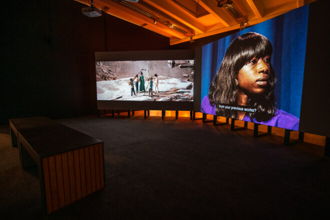 A pair of large projections in a dark gallery space. The left screen shows a film set and the right screen shows a panel show host.